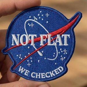 May include: Embroidered blue patch with white text reading "NOT FLAT" and "WE CHECKED". The patch features a red swoosh design and white stars, resembling a NASA logo. The patch is held by a hand.