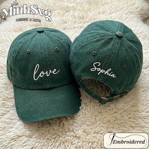 May include: Two dark green baseball caps. One cap has the word "love" embroidered in white, and the other has the name "Sophia" embroidered in white. The caps have adjustable straps and a curved brim. The image includes the text "MinhSvg DESIGN & ARTS" and "Embroidered".
