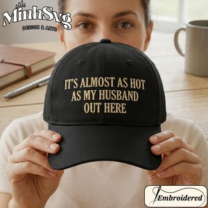 May include: A black baseball cap featuring gold embroidered text on the front that reads IT'S ALMOST AS HOT AS MY HUSBAND OUT HERE. The cap has a structured crown, a curved brim, and eyelets for ventilation, presenting a clean and simple design.