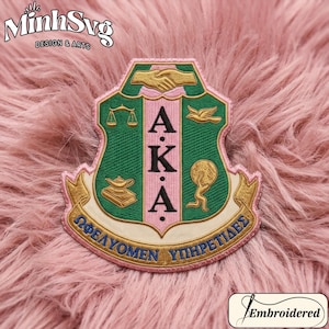 May include: Embroidered patch featuring the letters "AKA" in pink, set against a green shield design. The patch includes gold and pink accents, with Greek text at the bottom. The patch is on a pink, fluffy surface.
