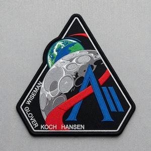 May include: Embroidered patch with a black, triangular border. The design features a stylized moon, Earth, and a blue and red graphic. The names "WISEMAN", "GLOVER", "KOCH", and "HANSEN" are printed on the patch.