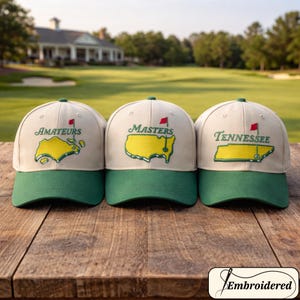 Op de afbeelding: Drie beige en groene baseballpetten met geborduurde ontwerpen. Elke pet heeft een ander ontwerp: "Amateurs", "Masters" en "Tennessee", met een gele kaartomtrek en een rode vlag.