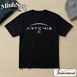 Puede incluir: Camiseta negra con el texto blanco "ARTEMIS II" en una fuente estilizada. El diseño está centrado en el pecho. La camiseta tiene cuello redondo y mangas cortas. La tela parece suave.
