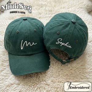 Puede incluir: Dos gorras de béisbol verde oscuro. Una gorra tiene la palabra "me" bordada en blanco en la parte delantera. La otra gorra tiene el nombre "Sophia" bordado en blanco en la parte trasera. Las gorras tienen una visera curva y una correa ajustable.