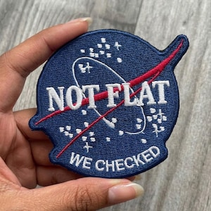 May include: A navy blue patch with white text that reads "NOT FLAT" and "WE CHECKED". The patch features a red diagonal line and a white outline of a shape, with white star-like shapes. The patch is round with a pointed end.