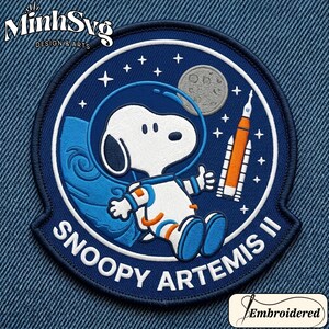 May include: Embroidered patch featuring Snoopy in an astronaut suit, with the text "Snoopy Artemis II". The patch is circular, with a dark blue background, white stars, a moon, and a rocket ship. The patch also includes the text "MinhSvg Design & Arts".