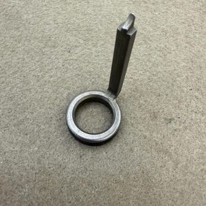 Könnte beinhalten: Ein silberfarbenes Metallwerkzeug mit kreisförmiger Basis und einem rechteckigen Griff, der sich nach oben erstreckt. Der Griff hat ein gekerbtes Ende. Das Werkzeug wird wahrscheinlich für einen bestimmten mechanischen oder handwerklichen Zweck verwendet.