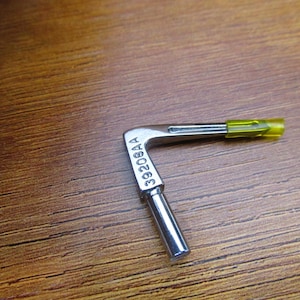May include: A silver-colored, L-shaped metal tool with a cylindrical handle and a yellow-green tip. The text "33208AA" is stamped on the tool. The tool is set against a brown wooden surface.