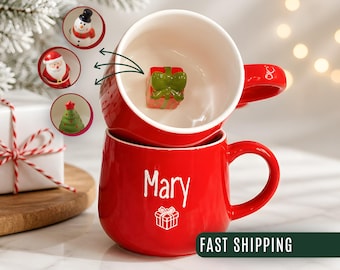 May include: Two stacked red ceramic mugs with white interiors. The top mug features a small, three-dimensional green and red gift box figurine inside. The bottom mug is personalized with the white text Mary and a small white gift box icon.