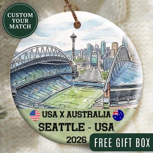 May include: A round ceramic ornament featuring a watercolor illustration of a stadium and the Seattle skyline with the Space Needle. Text reads USA X AUSTRALIA, SEATTLE - USA, and 2026, accompanied by the American and Australian national flags.