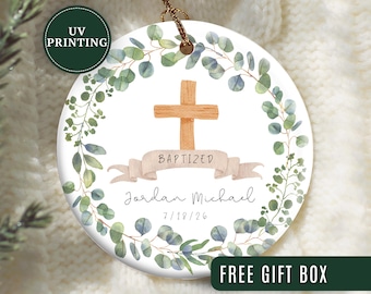 May include: A round, white ceramic ornament with a watercolor design. It features a wooden cross, the word "BAPTIZED" on a banner, and the name "Jordan Michael" with a date. The ornament is encircled by a green and blue eucalyptus wreath.