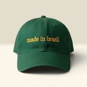 Può includere: Un cappellino da baseball verde scuro con la scritta "made in brasil" ricamata in filo dorato. Il cappellino ha una visiera curva e un bottone sulla parte superiore. Il cappellino è di colore verde scuro.
