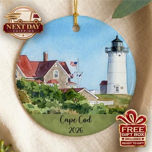 Könnte beinhalten: Ein rundes Keramikornament mit einer Aquarellillustration eines weißen Leuchtturms und eines Hauses mit rotem Dach. Die Szene zeigt eine kleine amerikanische Flagge, grünes Blattwerk und den Text Cape Cod 2026 am unteren Rand.