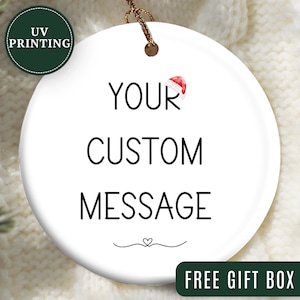 May include: A white, round ceramic ornament with the text "YOUR CUSTOM MESSAGE" in black. A small Santa hat graphic is above the word "YOUR". The ornament has a gold string for hanging and a green "UV PRINTING" logo.