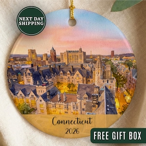 May include: A round ceramic ornament featuring a watercolor-style illustration of a university campus cityscape at dusk. The design includes stone buildings with dark roofs, warm glowing lights, and a sunset sky. The text Connecticut 2026 is printed below.