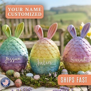 May include: Three egg-shaped figurines with rabbit ears and a textured scale pattern. Each figurine features a horizontal band with a personalised name: Jessica, Julia, or Sarah. The colours are a gradient of pastel green, yellow, pink, and purple.