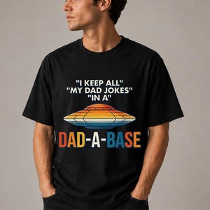 Op de afbeelding: Zwart T-shirt met de tekst "I KEEP ALL MY DAD JOKES IN A DAD-A-BASE" en een retro UFO-afbeelding in oranje, geel en blauw. Het shirt heeft een ronde hals.