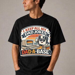 Puede incluir: Camiseta negra con un gráfico retro que presenta una computadora, un disquete y el texto "I KEEP ALL MY DAD JOKES DAD-A-BASE". El diseño tiene detalles en naranja, blanco y azul.