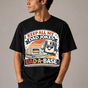 Puede incluir: Camiseta negra con un diseño gráfico retro que presenta un personaje de disquete con gafas de sol y un pulgar hacia arriba. El texto dice "I KEEP ALL MY DAD JOKES DAD-A-BASE" en una fuente vintage, con un ordenador y un diseño de puesta de sol.