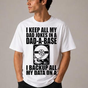 Könnte beinhalten: Weißes T-Shirt mit schwarzem Text und einer Festplattengrafik. Der Text lautet: "I KEEP ALL MY DAD JOKES IN A DAD-A-BASE. I BACKUP ALL MY DATA ON A". Das Shirt ist ein lässiger Stil.