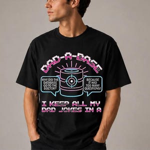 Op de afbeelding: Zwart T-shirt met een grafisch ontwerp. Het ontwerp bevat de woorden "DAD-A-BASE" in roze en wit, samen met een database-icoon en de tekst "I KEEP ALL MY DAD JOKES IN A". Het shirt bevat ook twee tekstballonnen met tekst.