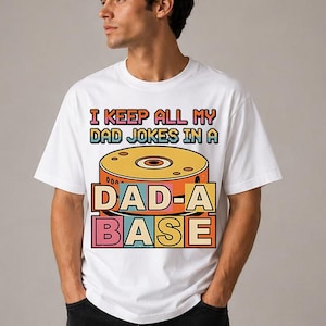 Puede incluir: Camiseta blanca con un diseño retro. La camiseta presenta el texto "I KEEP ALL MY DAD JOKES IN A DAD-A-BASE" en una fuente vintage colorida. El gráfico incluye una imagen estilizada de un CD o disco.