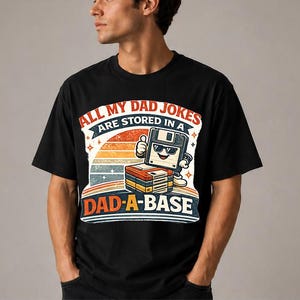 May include: Black t-shirt with a retro design. The graphic features a smiling floppy disk giving a thumbs up, with the text "ALL MY DAD JOKES ARE STORED IN A DAD-A-BASE" in a colorful, vintage style. The design includes a sunset and stars.