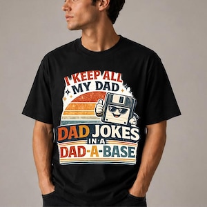 May include: Black t-shirt with a retro design featuring the text "I KEEP ALL MY DAD JOKES IN A DAD-A-BASE." The graphic includes a smiling floppy disk character wearing sunglasses, set against a sunset background.