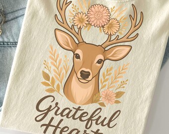Op de afbeelding: Crèmekleurig T-shirt met een hertenafbeelding, bloemaccenten en de tekst "Grateful Heart". De afbeelding bevat een lichtblauwe jeans, een gouden ketting en een beige hoed. Het T-shirt is gemaakt van zacht materiaal.