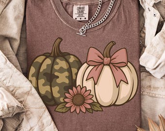 May include: A maroon Comfort Colors t-shirt featuring a fall-themed graphic. The design includes two pumpkins, one with a camouflage pattern and the other with a pink bow, along with a sunflower. A silver chain necklace and a tan jacket complete the look.