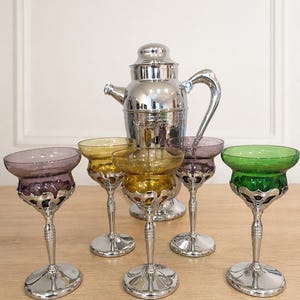 May include: A silver cocktail shaker with a handle and a set of five stemmed glasses. The glasses feature coloured bowls in purple, yellow, and green, each set into a decorative silver metal base with a matching silver stem and circular foot.