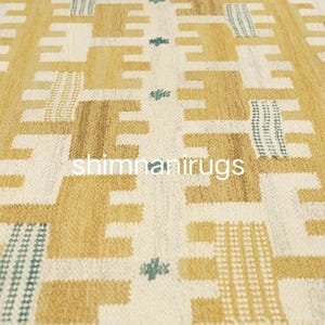 Op de afbeelding: Close-up van een geweven tapijt met een geometrisch patroon in tinten beige, bruin en gebroken wit. Het ontwerp bevat herhalende vormen en accenten van blauwgroen. De tekst "shimnanirugs" is zichtbaar.