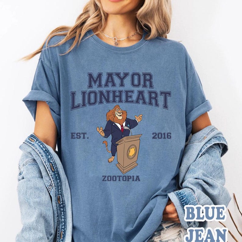 Puede incluir: Camiseta azul con un le&oacute;n de dibujos animados con traje en un podio. El texto "MAYOR LIONHEART" est&aacute; encima del le&oacute;n, con "EST. 2016" a ambos lados. La palabra "ZOOTOPIA" est&aacute; debajo del le&oacute;n.