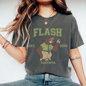 Puede incluir: Camiseta gris oscuro con un perezoso de dibujos animados de Zootopia. La camiseta tiene la palabra "FLASH" en letras verdes en bloque sobre el perezoso, con "EST. 2016" y "ZOOTOPIA" impresos debajo.