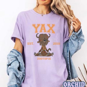 Puede incluir: Una camiseta de color lila claro con un personaje de yak de dibujos animados de Zootopia. El dise&ntilde;o incluye el texto "YAX" en letras naranjas en bloque sobre el yak, con "EST. 2016" a cada lado. La palabra "ZOOTOPIA" est&aacute; debajo.