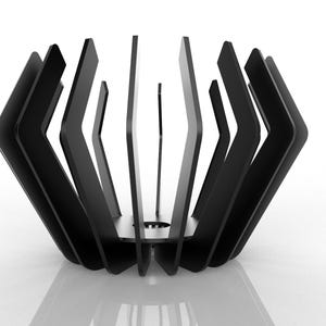 Puede incluir: Un objeto decorativo geométrico negro con una serie de láminas angulares verticales dispuestas en un patrón circular. Las láminas están espaciadas uniformemente y crean una estructura similar a una jaula. El objeto se encuentra sobre una superficie blanca.
