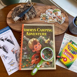 May include: A spiral-bound book titled Tommy's Camping Adventure, a magnifying glass with a green handle, a Project FeederWatch bird guide, a box of 8 Crayola crayons, a blue cup with crayons, and several nature-themed stickers on a wooden board.
