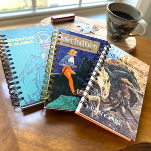 May include: Three spiral-bound notebooks featuring vintage book cover art. One is light blue with text reading Adventure in Alaska, another shows a person riding a black horse titled Timber Trail Riders, and the third depicts horses titled Fury and the Mustangs.