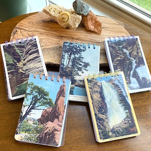 May include: Five small spiral-bound notebooks with vintage landscape images. The notebooks feature images of waterfalls, rock formations, and trees. The notebooks are arranged on a wooden surface.