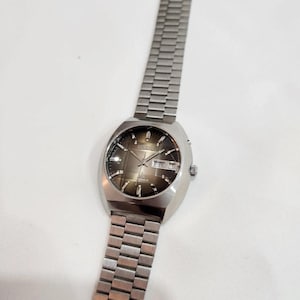 以下が含まれることがあります： 長方形の文字盤と金属バンドが付いた、ヴィンテージのシルバーカラーの腕時計。文字盤は濃い茶色で、曜日と日付を示す白い文字があります。バンドは、小さな長方形の金属リンクでできています。