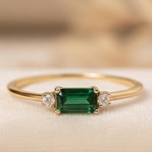 Puede incluir: Un anillo de oro con una esmeralda verde rectangular, flanqueada por dos piedras preciosas redondas y transparentes más pequeñas. El anillo está sobre una superficie de terciopelo blanco, con un fondo suave y borroso.