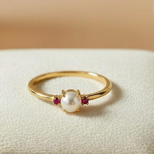Könnte beinhalten: Ein goldener Ring mit einer zentralen weißen Perle und zwei kleinen roten Edelsteinen. Der Ring liegt auf einer weißen Samtoberfläche und zeigt sein zartes Design. Der Ring hat ein dünnes und elegantes Band.
