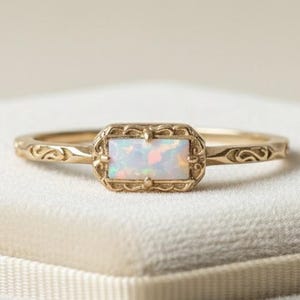 Könnte beinhalten: Ein goldener Ring mit einem Opalstein. Der rechteckige Opal ist in einem dekorativen Rahmen gefasst. Das Band weist ein eingraviertes Design auf. Der Ring wird auf einer weißen Samtoberfläche präsentiert.