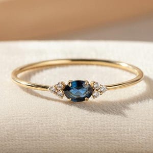 Op de afbeelding: Een delicate gouden ring met een diepblauwe ovale edelsteen in het midden, geflankeerd door kleinere, sprankelende accentstenen. De ring ligt op een zacht, crèmekleurig oppervlak, wat het elegante ontwerp benadrukt. Een fijn sieraad.