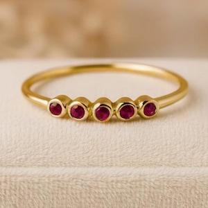 Puede incluir: Un delicado anillo de oro con cinco pequeñas gemas rojas redondas dispuestas en fila. El anillo tiene una banda fina y un acabado pulido y brillante. El anillo se muestra sobre una superficie suave y clara.