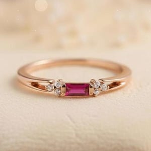 May include: Rose gold ring with a rectangular, deep pink gemstone. The ring is adorned with small, round, clear gemstones on either side of the central stone. The band has a delicate, split design. The ring is presented on a soft, off-white surface.