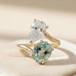 Puede incluir: Un anillo de oro con dos piedras preciosas en forma de pera, una transparente y otra verde. El anillo se exhibe sobre una superficie de terciopelo beige, resaltando el diseño y el contraste de color. Las piedras miden aproximadamente 1,3 cm de largo.