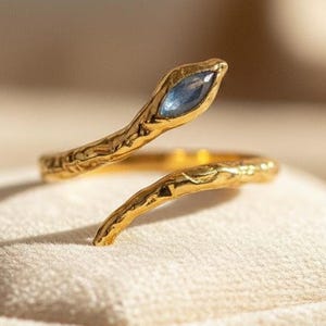 May include: A gold snake ring featuring a blue gemstone. The ring has a textured, organic design, with the snake's head and tail wrapping around the finger. The ring is displayed on a cream-coloured surface.