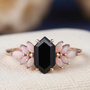 May include: Rose gold ring with a black, hexagonal central stone, surrounded by light pink opal stones. The ring is displayed on a cream-coloured surface. A striking piece of jewellery with a contrast of colours and textures.