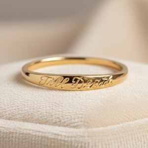 May include: A gold-colored ring with the inscription "Till Death" engraved on the band. The ring is displayed on a white velvet surface, showcasing its polished finish and elegant design. A classic piece of jewelry.